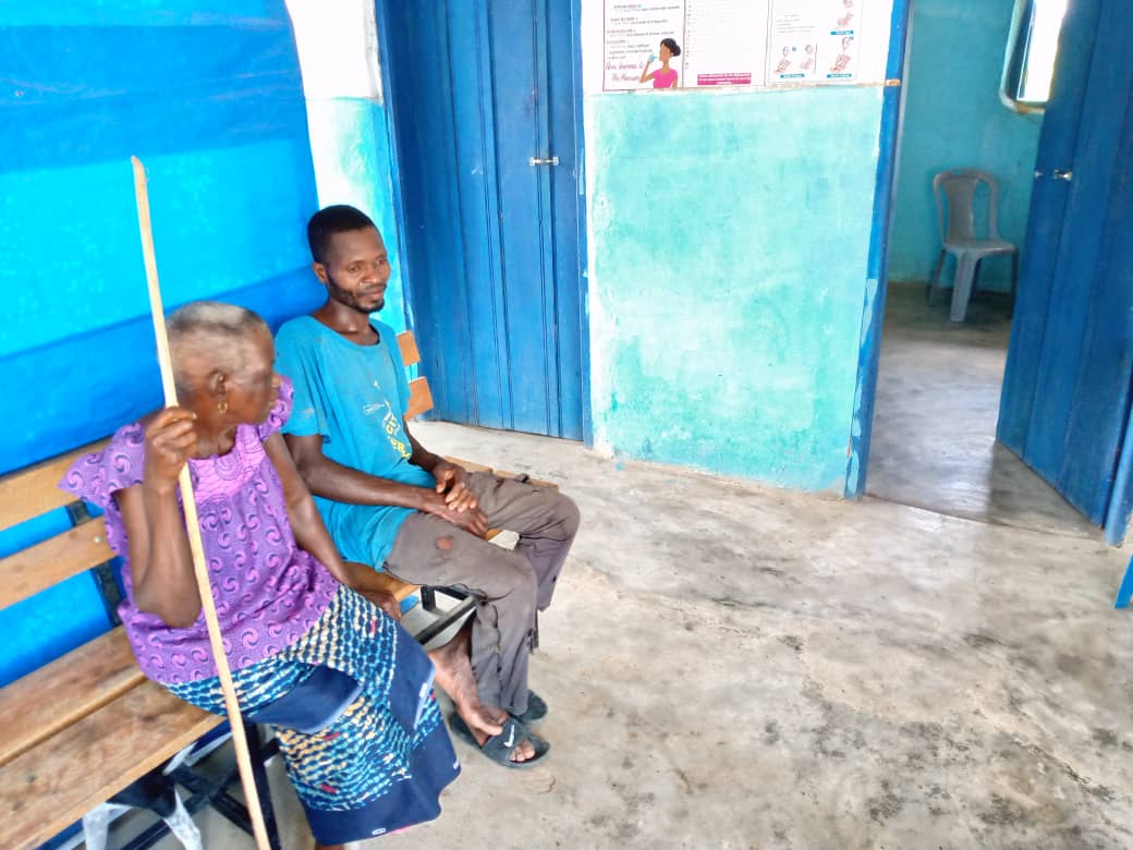 An elderly mother & son sitting on a well-crafted bench as they wait for wellness service.