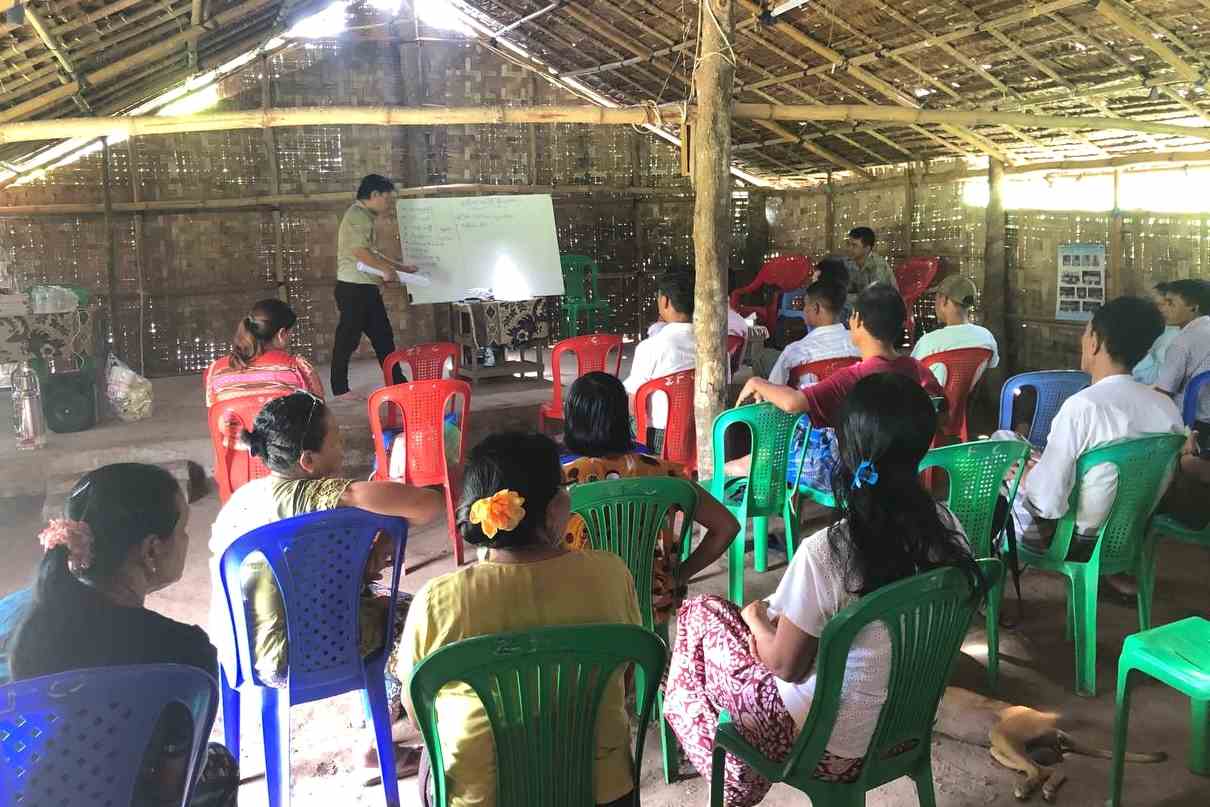 A near-filled, Myanmar TCD meeting happening as the teacher is explaining what's on the whiteboard.