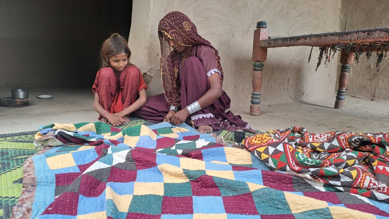 A Pakistan woman sewing a bed sheet as a Pakistan girl watches her.