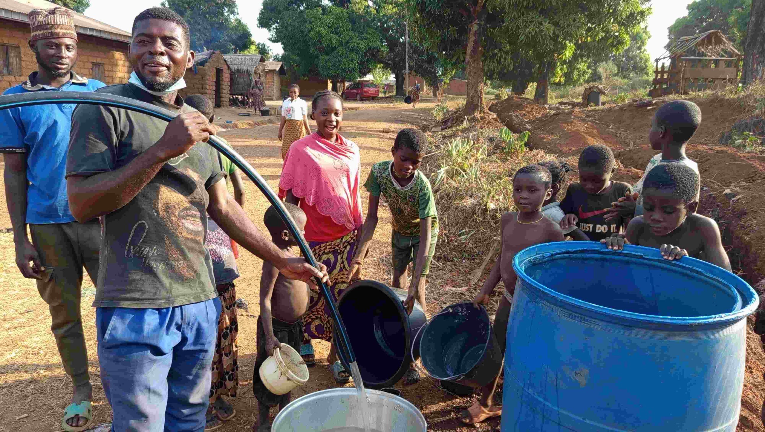 A Cameroon villager pouring water in a large barrel as they village children watch.